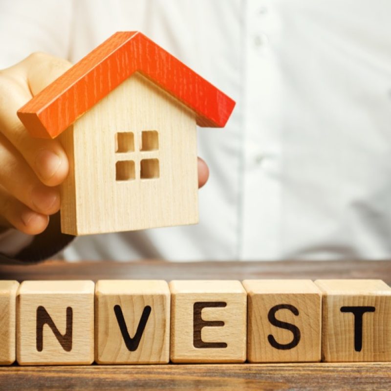 Wooden blocks with the word Invest and house in the hands of a businessman. The concept of investing in real estate construction. Effective investment. Buying apartments. Business and finance